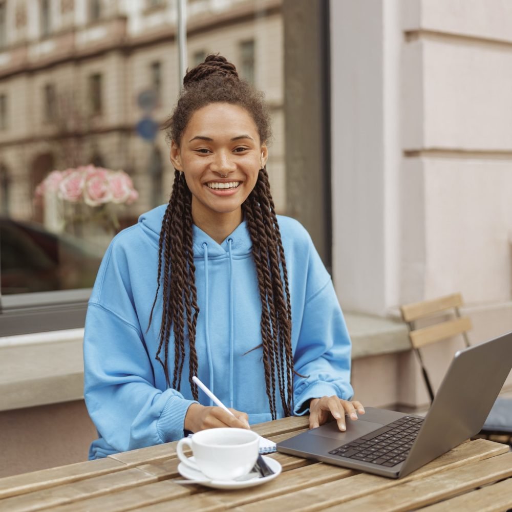 happy-pretty-young-woman-with-braids-and-piercing-at-laptop-smiling-and-sitting-at-cafe-outdoor-.jpg